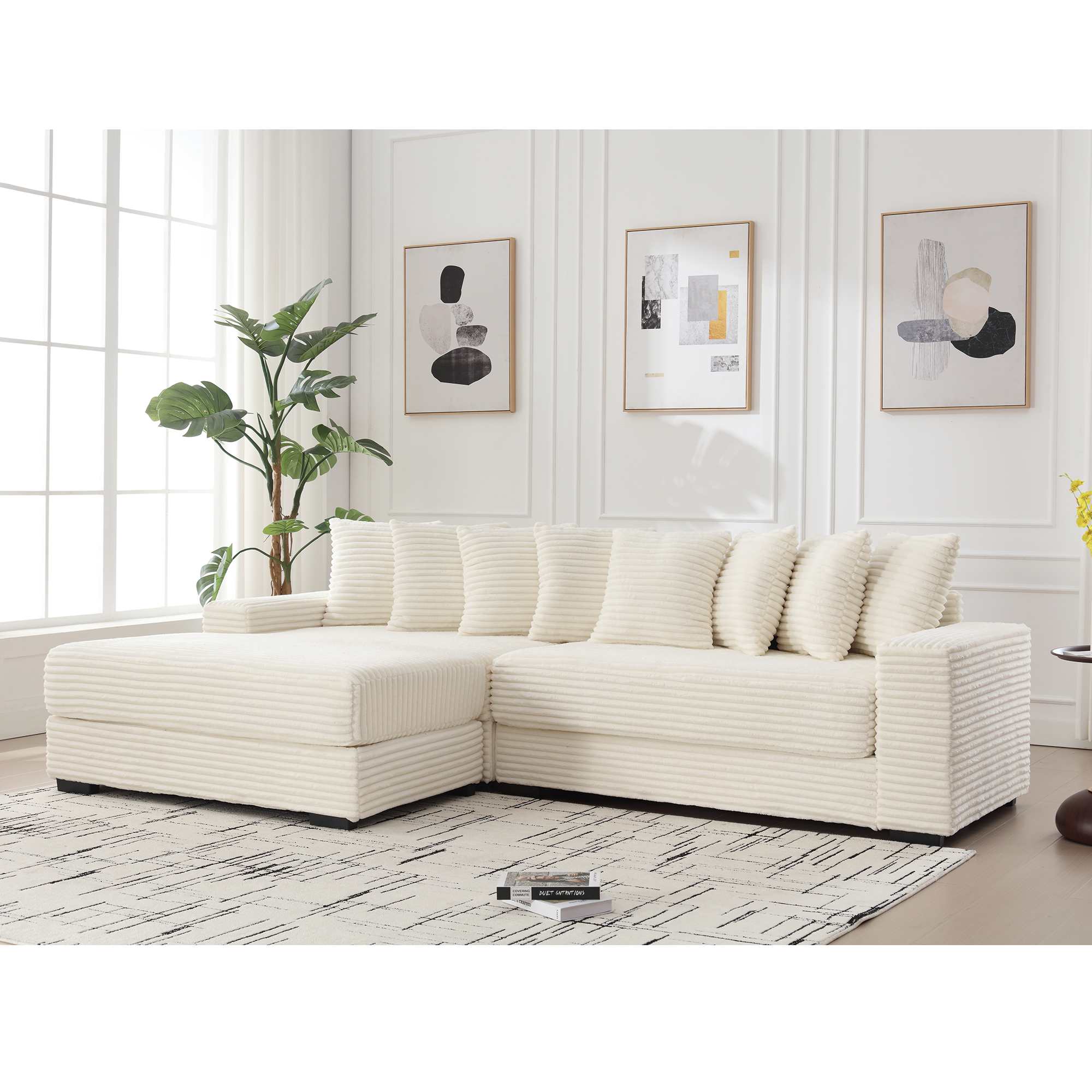 Cream corduroy L-shaped sofa with left chaise daybed and armrests, eight matching throw pillows, placed on patterned rug in bright living room with abstract wall art and large window.