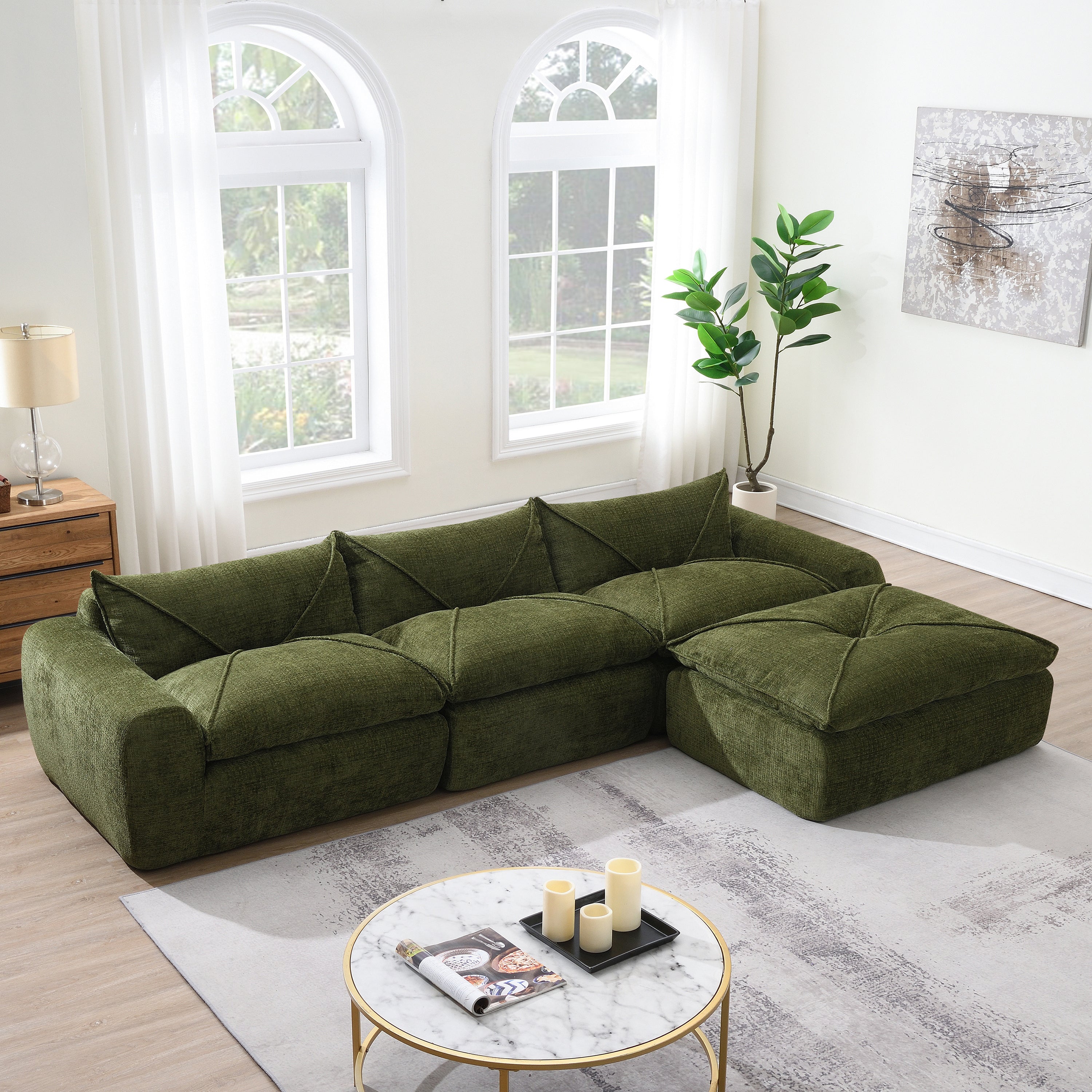 Green sectional sofa with integrated backrest and seat cushions placed in a bright living room with large arched windows, sheer white curtains, a round marble coffee table with candles, and a potted plant.