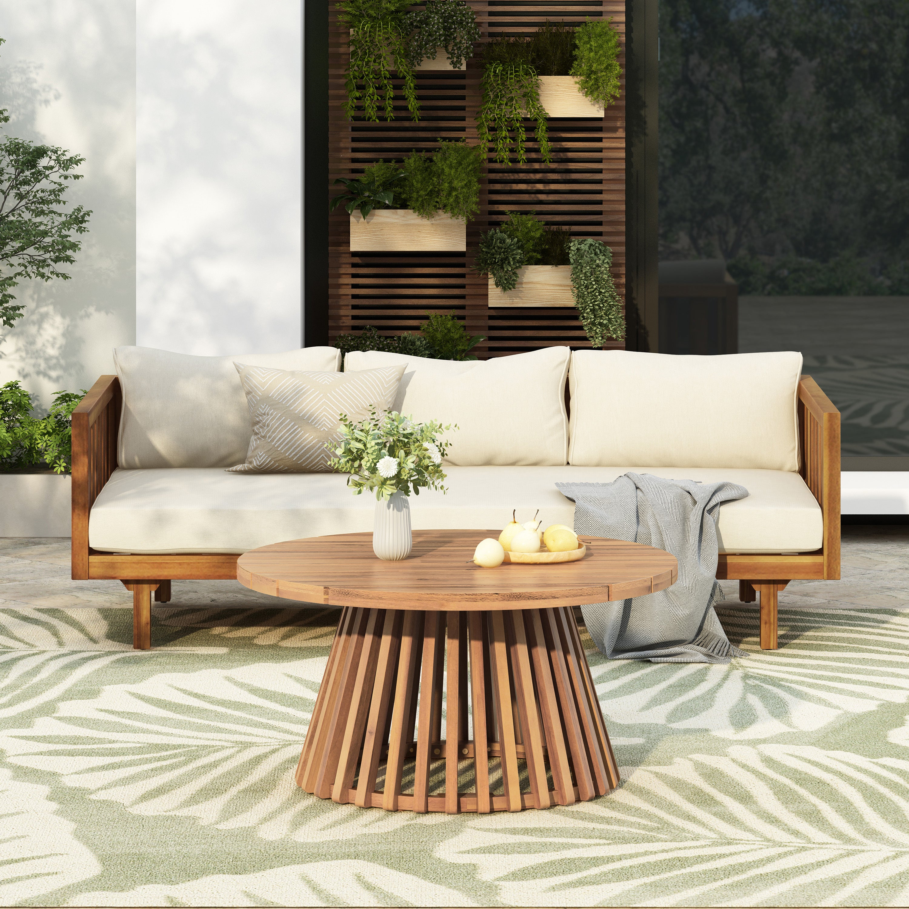 Outdoor patio furniture set with a three-seater daybed featuring light cushions and a round acacia wood table on a patterned outdoor rug, surrounded by greenery in a garden setting.