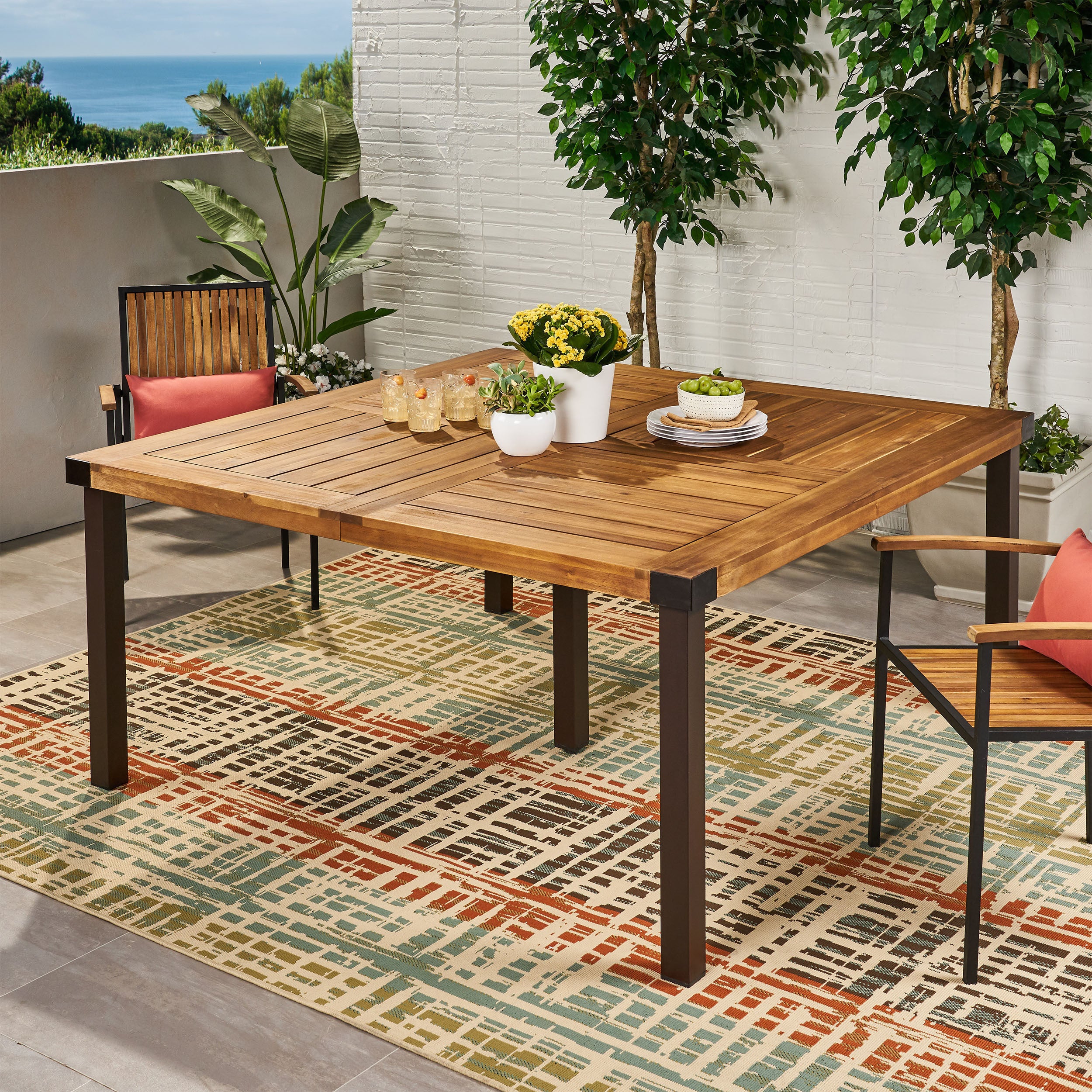 Square teak outdoor table with dark metal legs on a patterned rug, surrounded by wooden chairs with coral cushions, set in a garden patio with plants and ocean view, Garden & Outdoor collection.