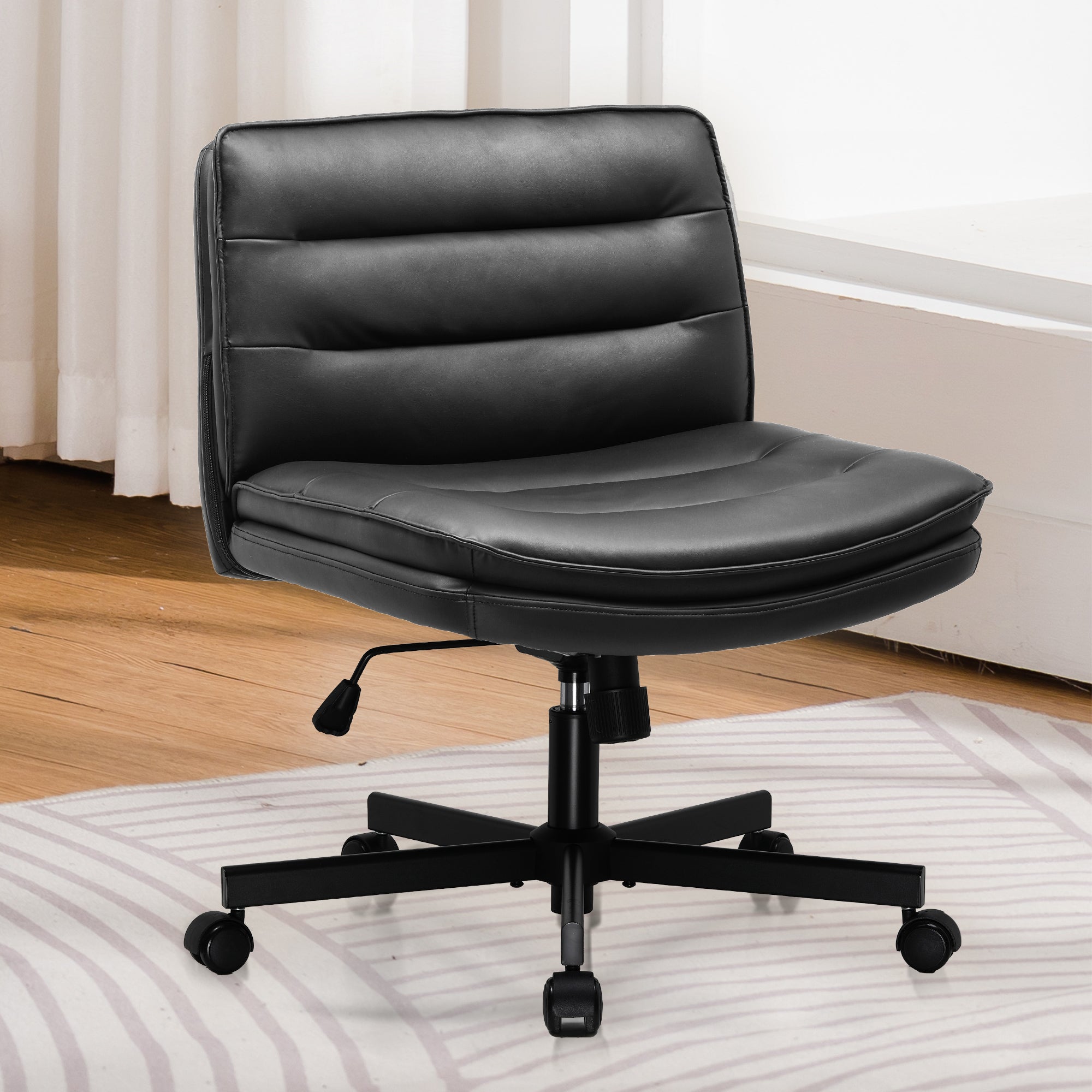 Black ergonomic armless office chair with adjustable height, padded seat and backrest on caster wheels, set on a wooden floor near a white desk and curtains.