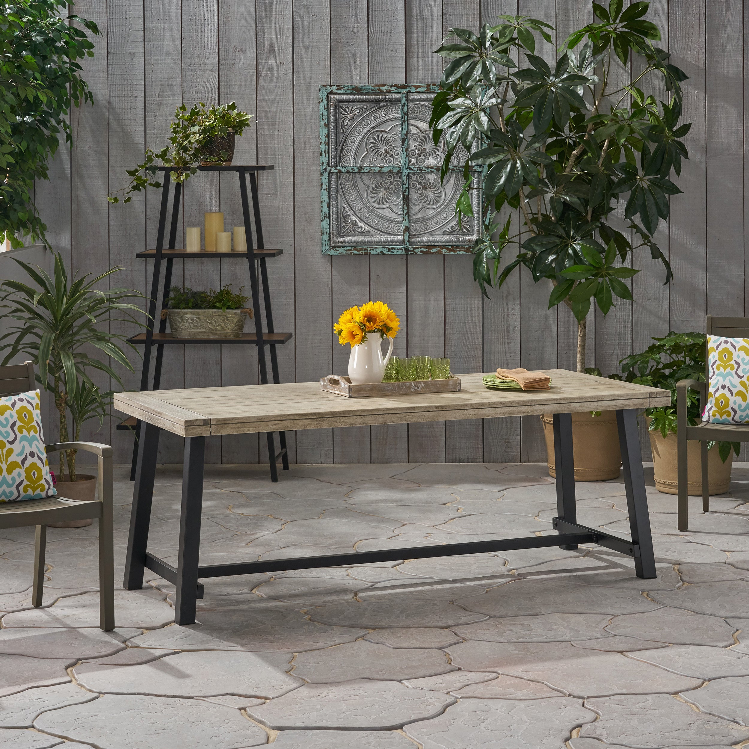 Wood outdoor patio table with grey wood top and black metal legs on stone patio, surrounded by garden plants and chairs with colorful cushions from Garden & Outdoor furniture collection.