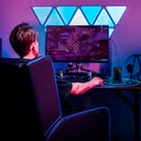 Man sitting in a black recliner chair with blue accent lighting, playing a video game on a widescreen monitor with triangular LED wall lights above, in an office and gaming setup