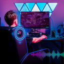 Man sitting in ergonomic office chair with massage and Bluetooth speakers, playing video games at desktop setup with colorful LED wall panels and soundwave visualization graphic.