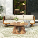 Outdoor patio furniture set with a three-seater daybed featuring light cushions and a round acacia wood table on a patterned outdoor rug, surrounded by greenery in a garden setting.