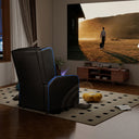 Black ergonomic recliner chair with blue LED accents positioned in a modern living room with a large projected screen, wooden cabinet, and patterned rug in an office and game furniture setting.