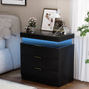 Black nightstand with drawers and sliding top featuring blue LED lights, decorated with a small white table lamp, framed art, books, and a flower vase in a bedroom setting.
