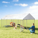 A large outdoor metal dog kennel with a white canopy roof is set up on grass, containing a dog house, elevated dog bed, and ramp, while a woman kneels near the open gate.