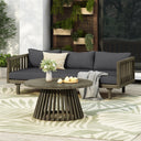 Outdoor patio scene featuring a dark grey cushioned three-seater daybed with acacia wood frame, round coffee table, decorative pillows, throw blanket, and potted plants wall display in garden and outdoor setting.