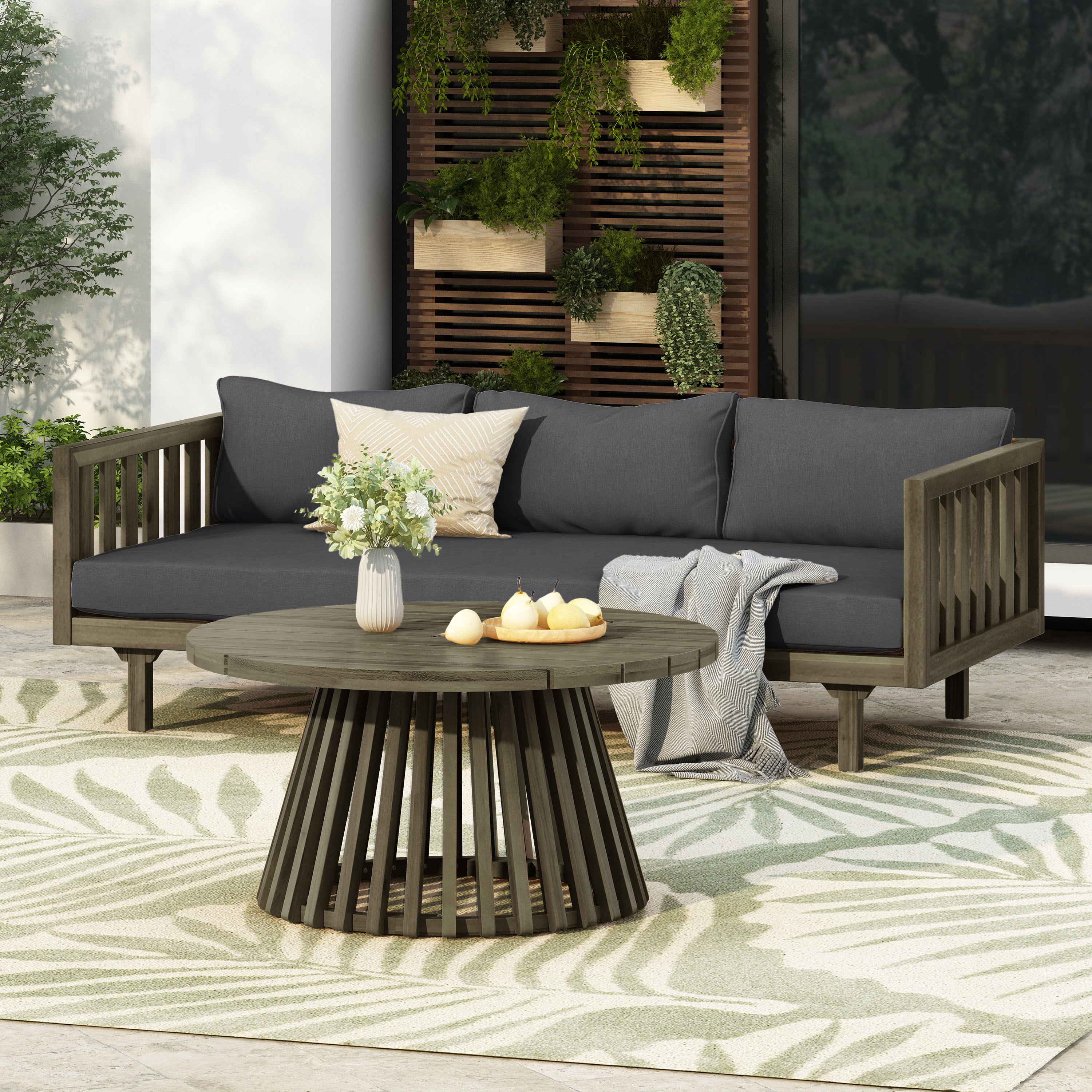 Outdoor patio scene featuring a dark grey cushioned three-seater daybed with acacia wood frame, round coffee table, decorative pillows, throw blanket, and potted plants wall display in garden and outdoor setting.